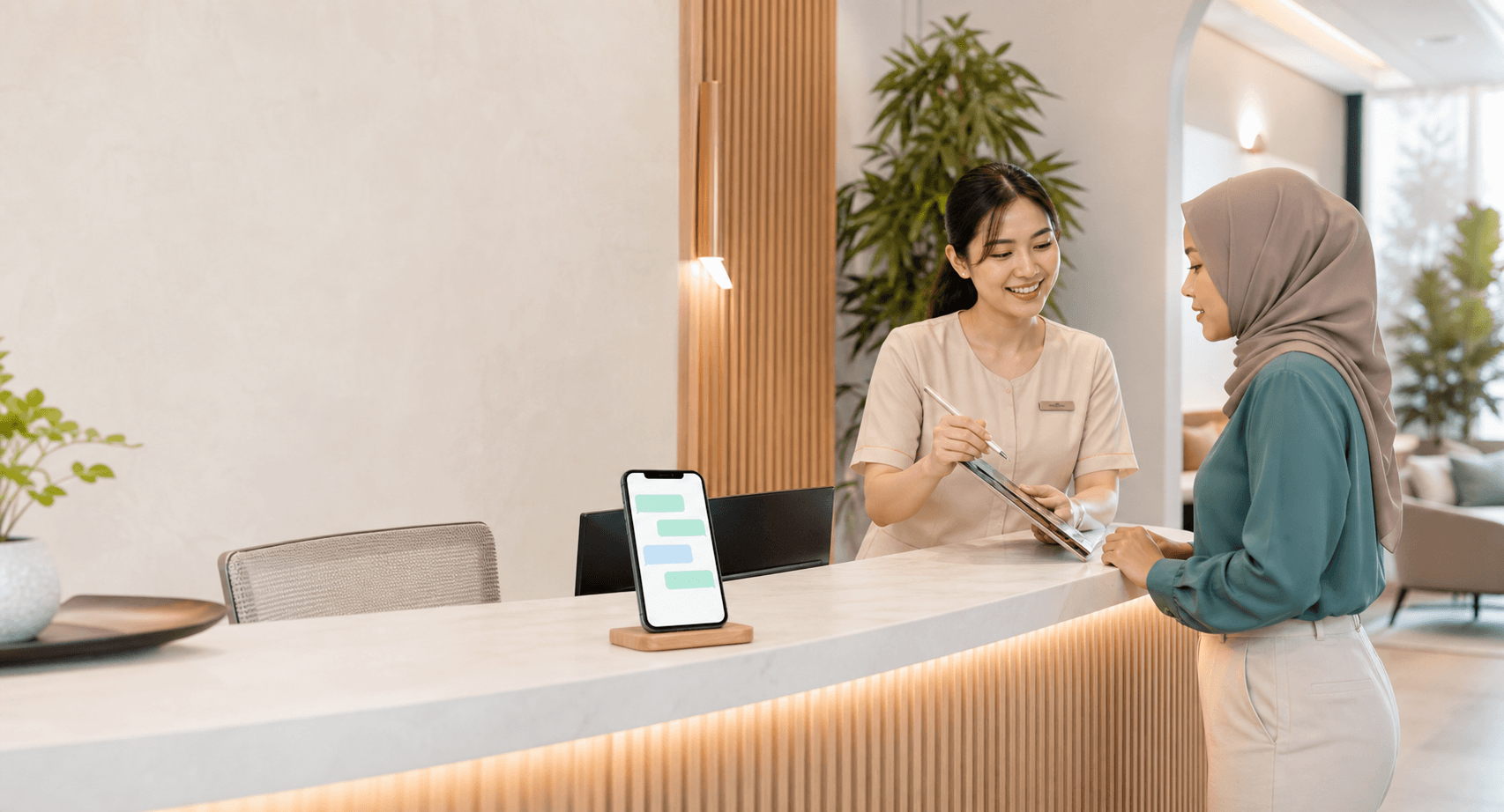 A Malaysian wellness receptionist helping a customer while a phone displays simple message bubbles